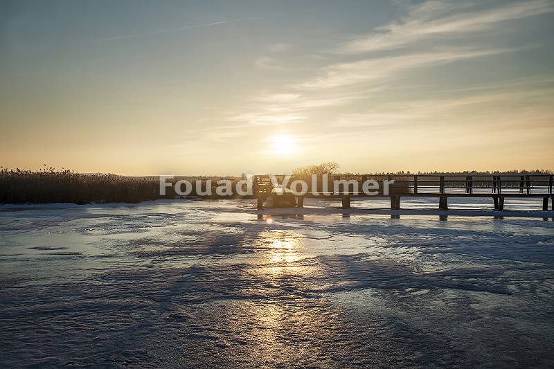 Gefrorener See mit Holzsteg im Sonnenuntergang